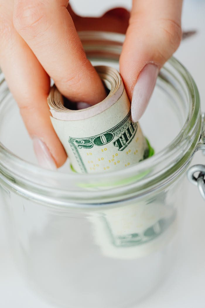 our-services-1 Hand placing rolled US dollars into a glass jar, representing savings and financial planning.